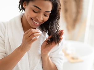 非洲裔美国妇女在家里的现代浴室中对受损干燥的头发使用血清。
