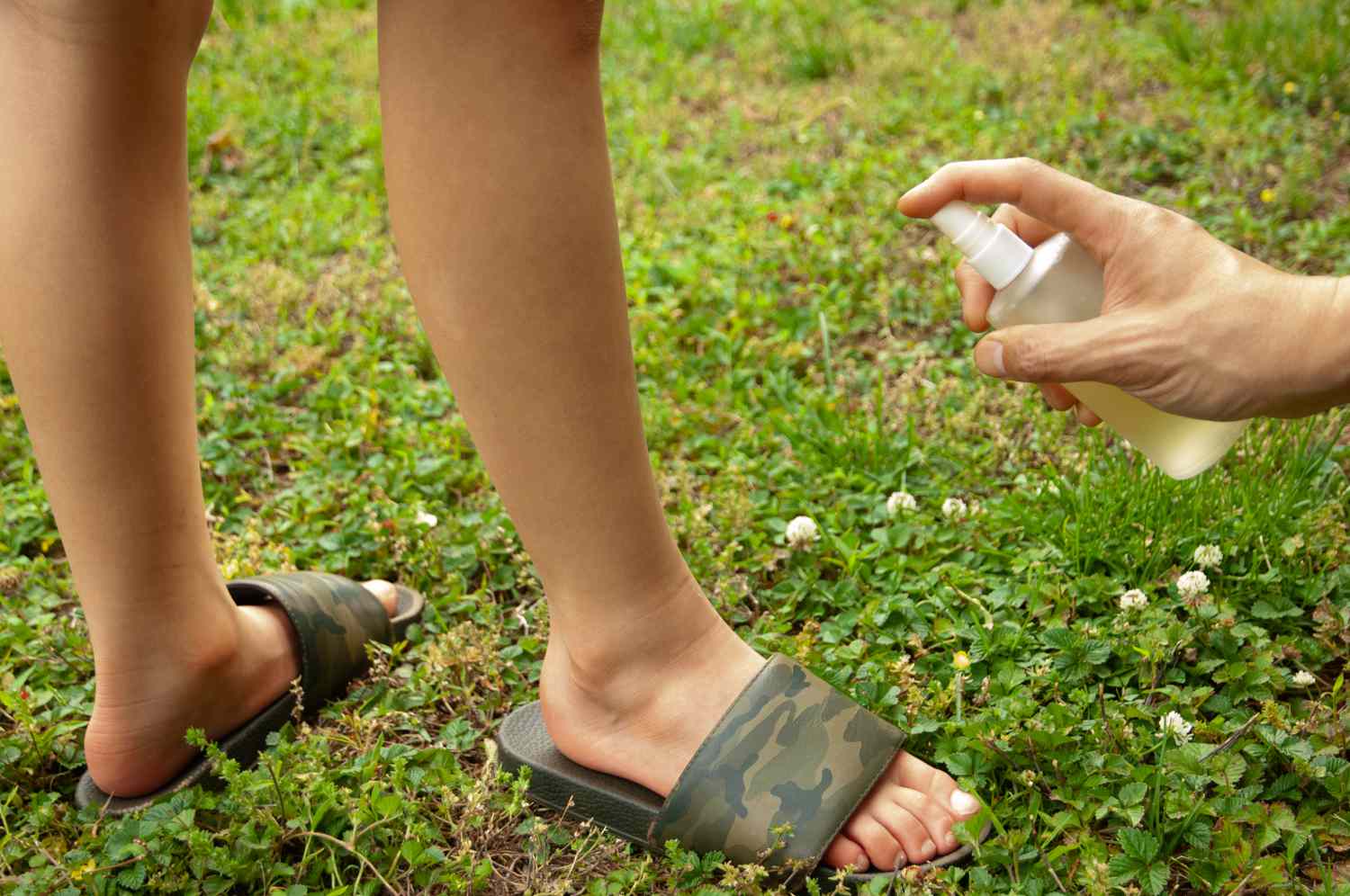 手在外面的草地上喷洒纯天然的驱虫剂在孩子的腿上＂width=