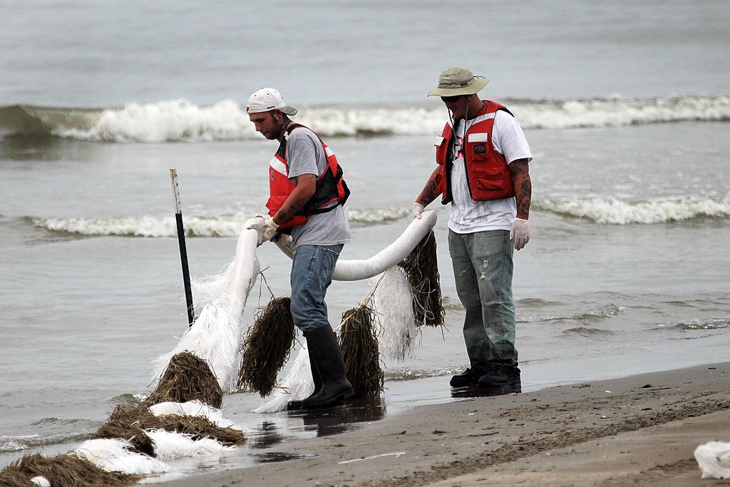 红色背心的油清洁工人试图使残留物从深水地平线漏油中泄露到路易斯安那州大岛的海滩上时，将吸收性材料放在水边。