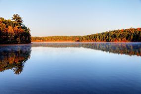 秋天在沃尔登池塘上有完美平坦的蓝色水，周围有五颜六色的树木，上面有浅蓝色的天空