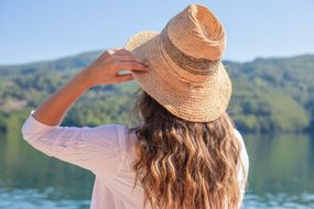 在炎热的夏天，一个女人戴着一顶大草帽站在湖边＂width=