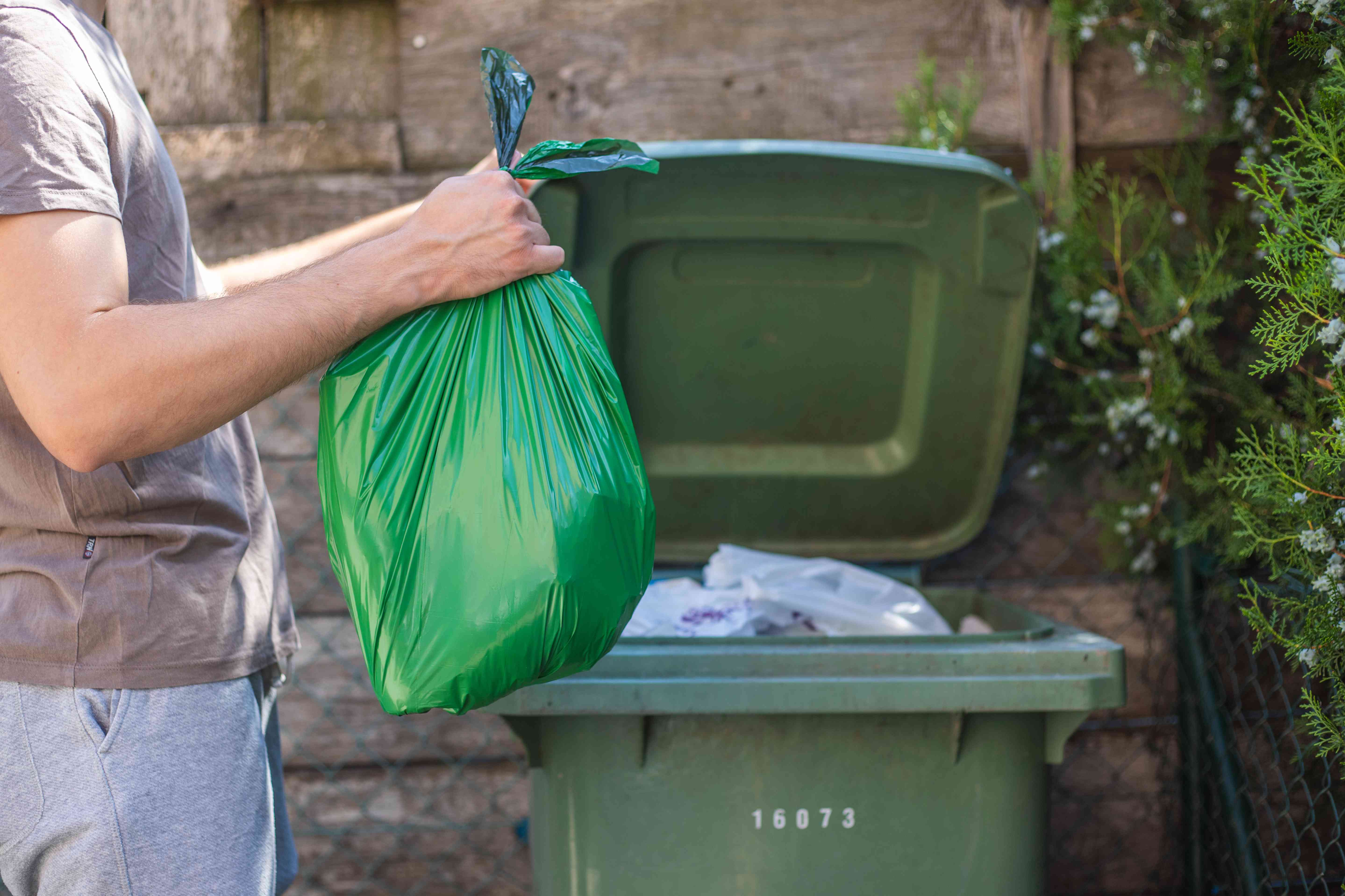 guy puts green plastic trash bag into household trash bin outside
