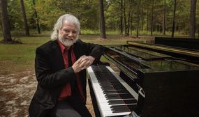 音乐家查克·莱维尔（Chuck Leavell）在树林里的钢琴上。“width=