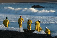 溢油清洁工人穿着黄色危险品齿轮和硬帽沿海滩铲石油污染的沙子，前景上有透明的塑料袋。