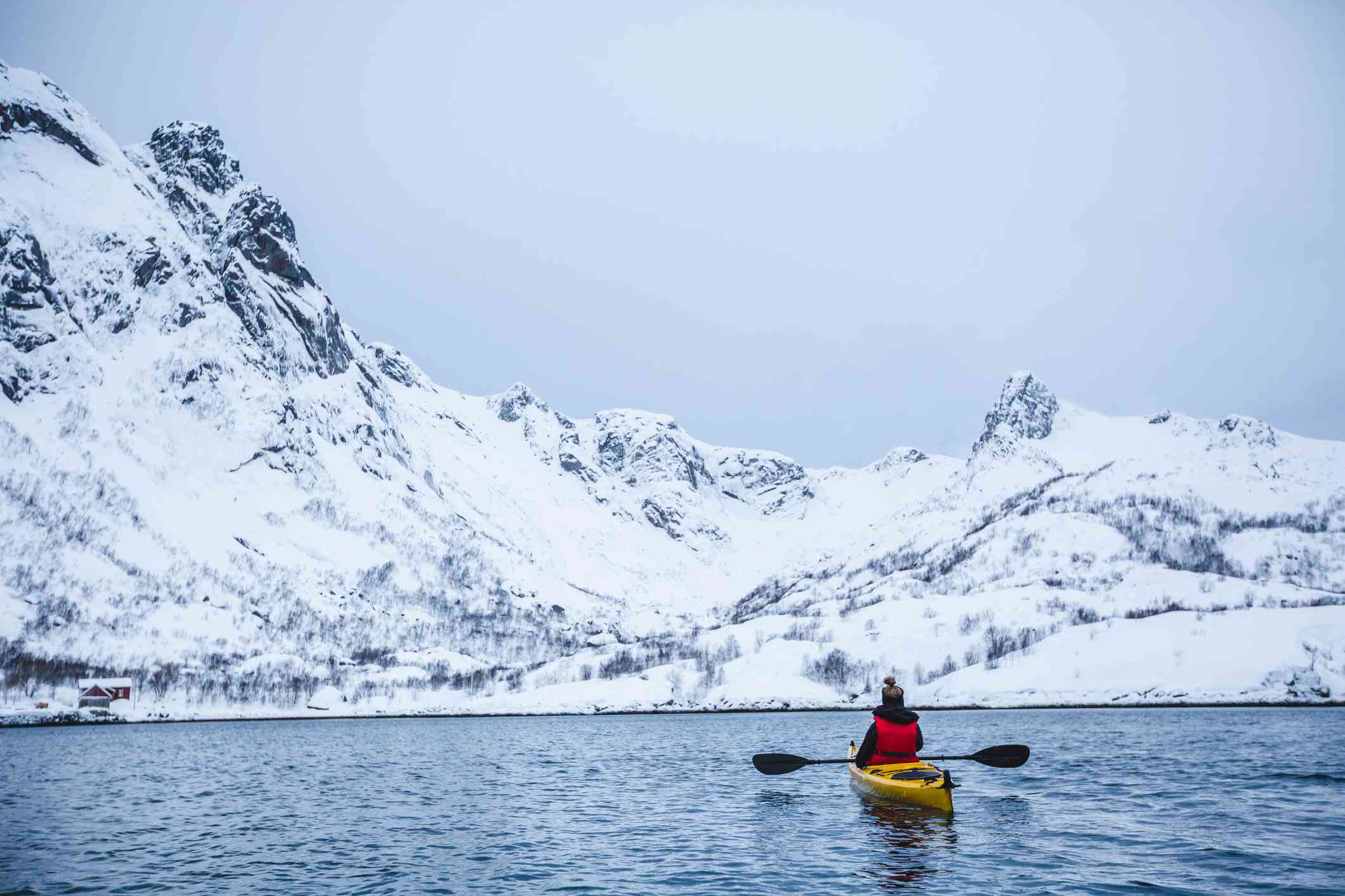 在挪威，一名划着黄色皮艇的单人皮艇手，桨向外伸展，面对着被雪覆盖的峡湾
