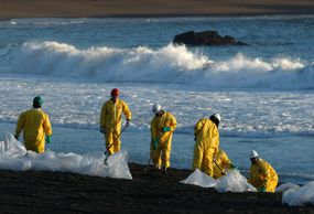 油污清理工人身穿黄色防护装备，头戴安全帽，沿着海滩铲被油污污染的沙子，前景是透明的塑料袋。