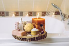 家用水疗产品木盘托盘:肥皂、浴炸弹,芳香浴盐,和按摩精油,蜡烛燃烧,卷起的毛巾在浴室的浴缸,水运行。舒适放松的概念。”width=