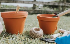 两个新鲜，新的，干净的赤土陶器罐在一个花园里，在篱笆前面。植物，绳子和前景中的各种其他园艺设备。“width=