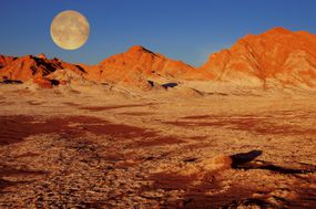 在智利的阿塔卡马沙漠中，夕阳下的一轮满月映照着红红的山脉。