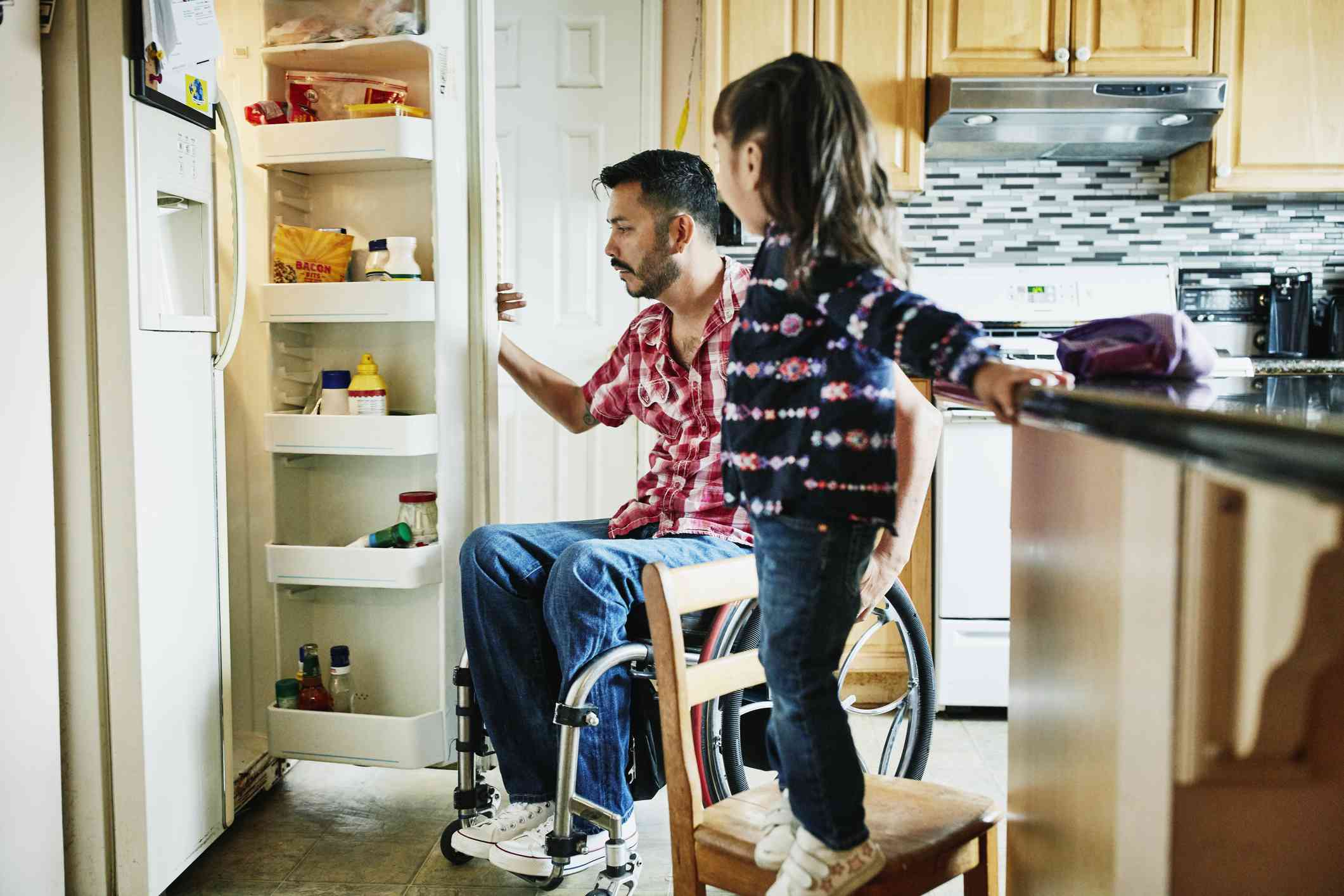 坐在轮椅上的父亲在厨房的冰箱里寻找，而女儿则站在椅子上看着＂width=