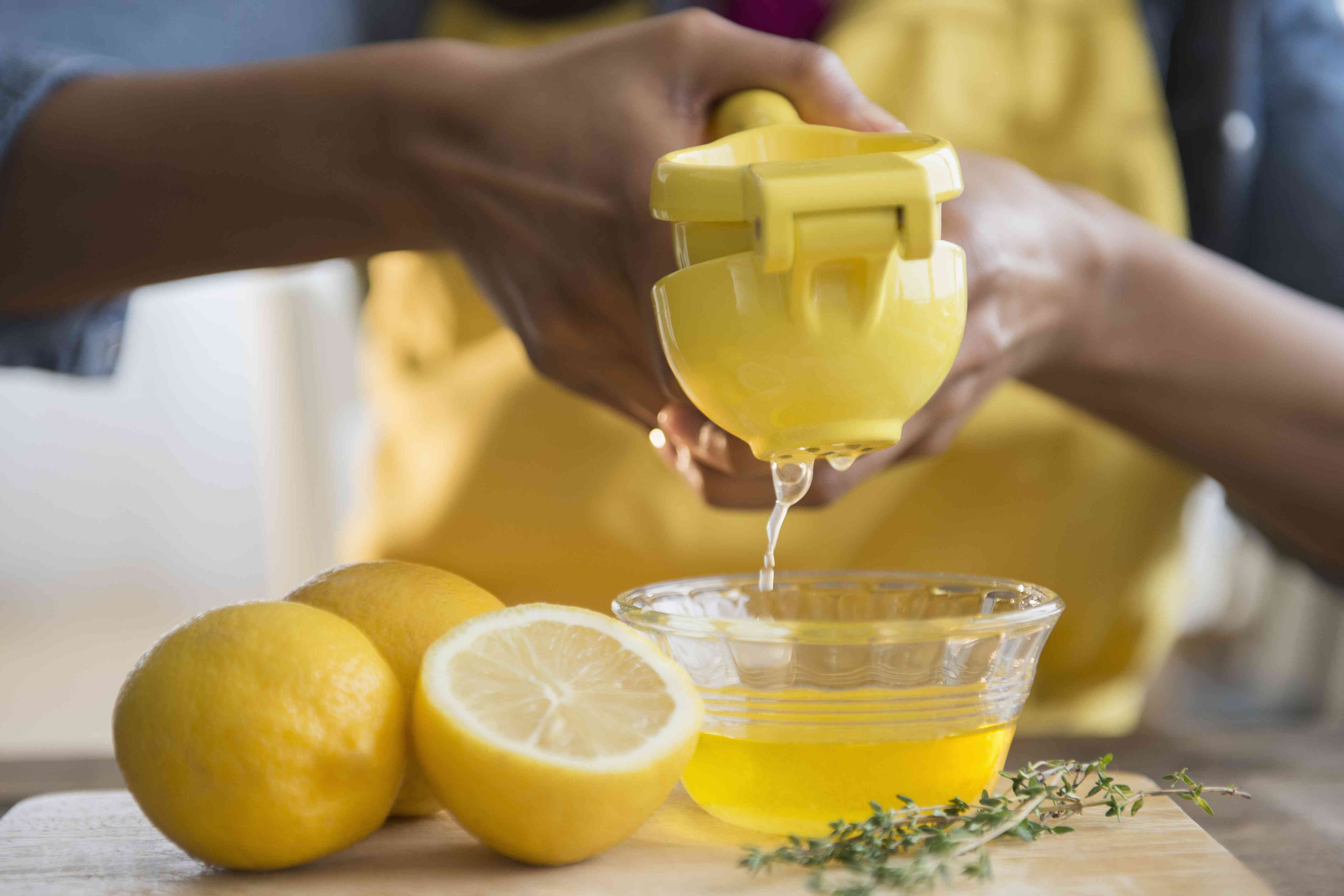 有人在一个小碗上使用金属柠檬汁挤压。更多的柠檬坐在旁边的工作台上，靠近百里香小树枝“width=