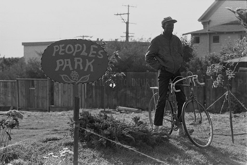加州伯克利，一名男子骑着自行车站在人民公园的一块标志牌旁。＂width=