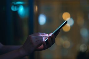 女性在黑暗中使用智能手机的手特写，在城市灯光的衬托下＂width=