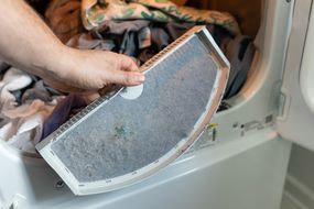 手从烘干机中抽出棉绒过滤器，显示出衣服棉绒的完整灰色衬里