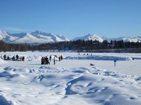 在一个蓝天的下午，雪山在背景中若隐若现，堆雪让位给一个冰冻的湖泊，湖面上有数十名滑冰运动员