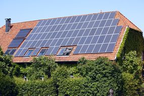 被绿色植物包围的房子的屋顶上的太阳能电池板。”width=