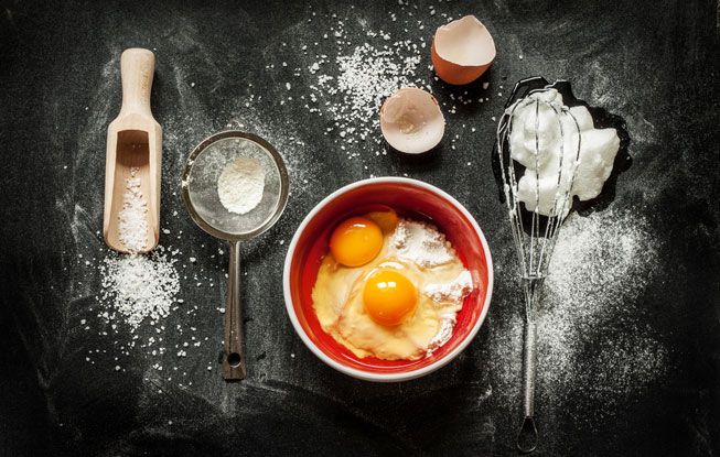 一碗鸡蛋，一些面粉和一个打蛋器放在黑色的桌子上