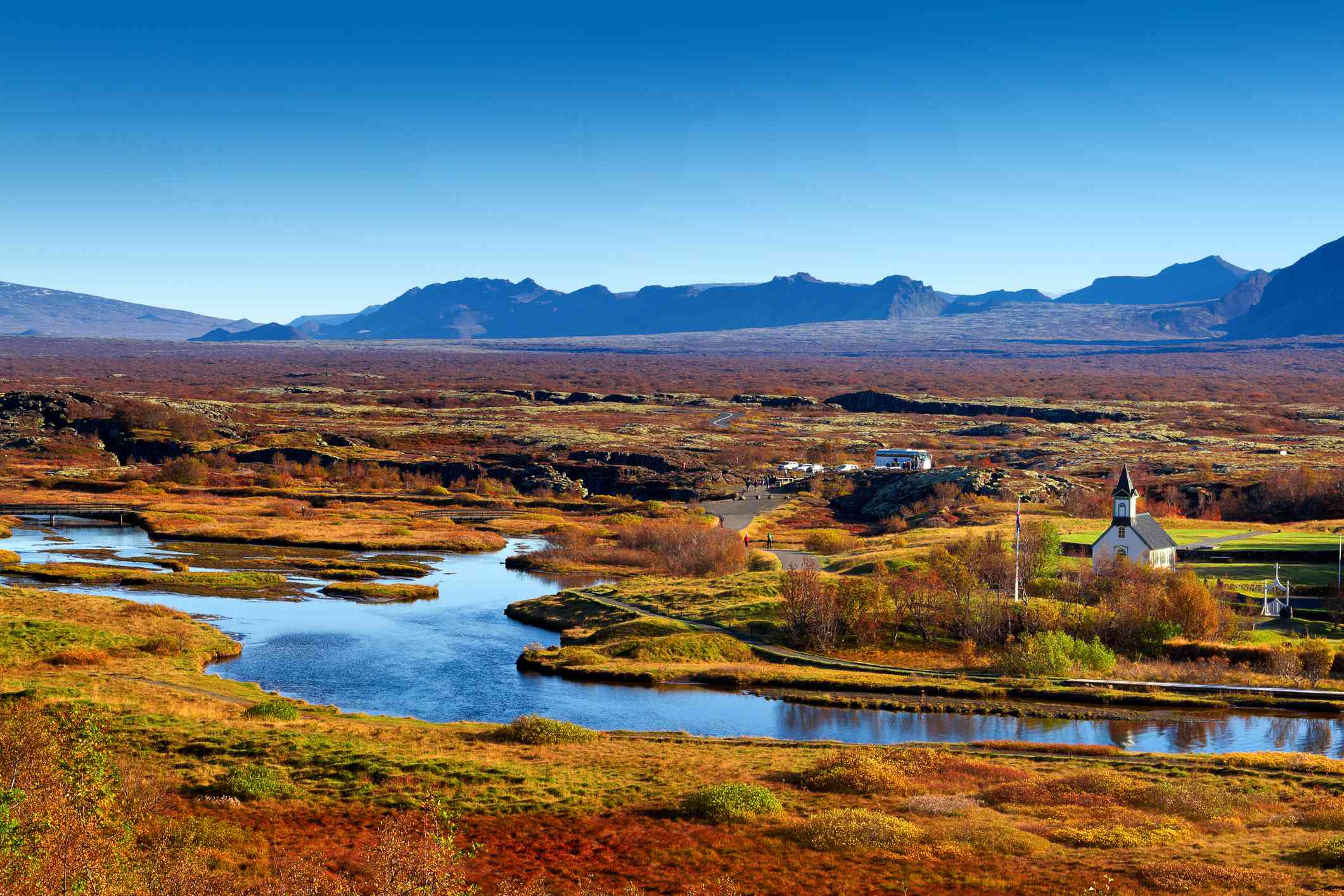 冰岛辛韦利尔国家公园奥克拉拉河畔的辛瓦拉基亚教堂，河两岸有深浅不一的红色、绿色和橙色的树木和植物，远处是群山和明亮的蓝天