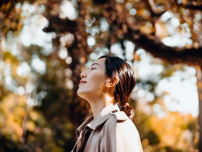 年轻的亚洲女子在公园散步的肖像，在美丽的秋日户外享受温暖的阳光，闭着眼睛呼吸新鲜空气。在自然的枫树下放松