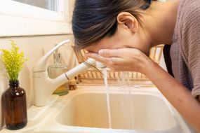 女人靠在白色的水槽上，用水和天然洗面奶洗脸”width=