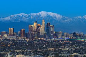 圣盖博山国家纪念碑的白雪覆盖的山峰超越洛杉矶市中心的天际线”width=