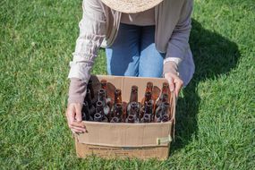 戴草帽的人蹲在草地上，手里拿着一个装着用过的棕色玻璃瓶的纸箱