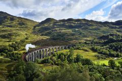格伦芬南高架桥俯瞰苏格兰高地郁郁葱葱的绿色山丘，詹姆斯党人蒸汽火车在蓝天白云下驶过