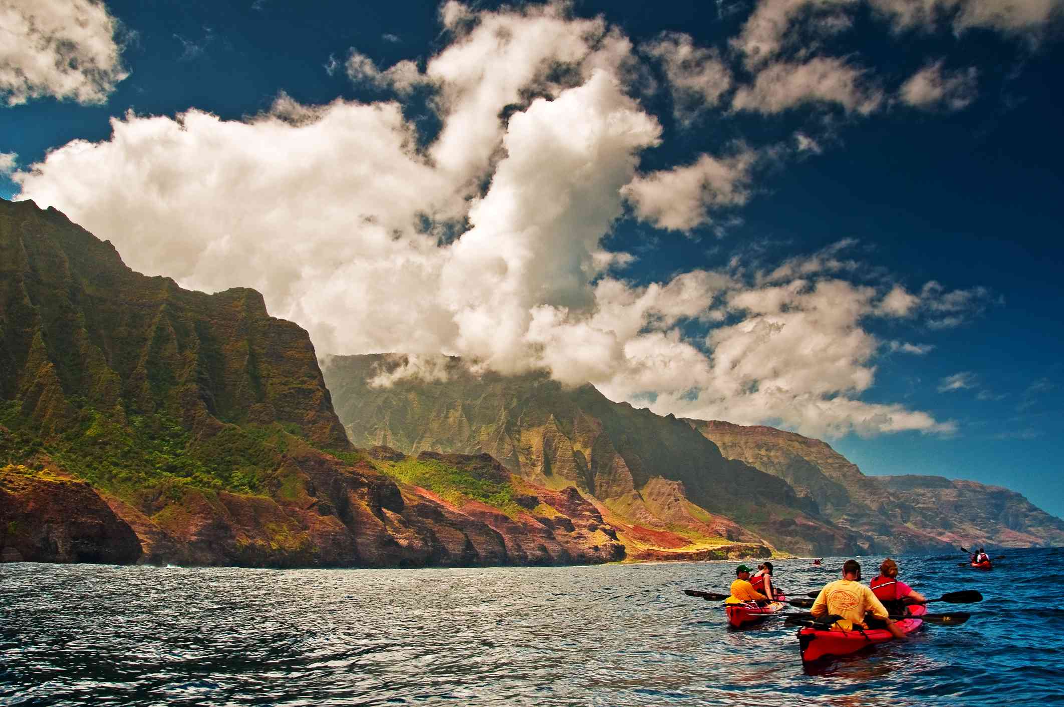 两对皮划艇在考艾岛纳帕利海岸附近的水域里，旁边是被绿色植物覆盖的平坦岩石，蓝天和白色的翻滚的云