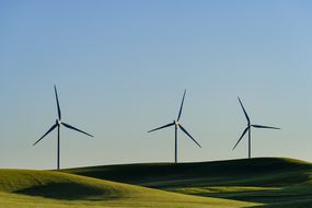 在起伏的山丘和蓝天的景观上，三个风力涡轮机。“width=