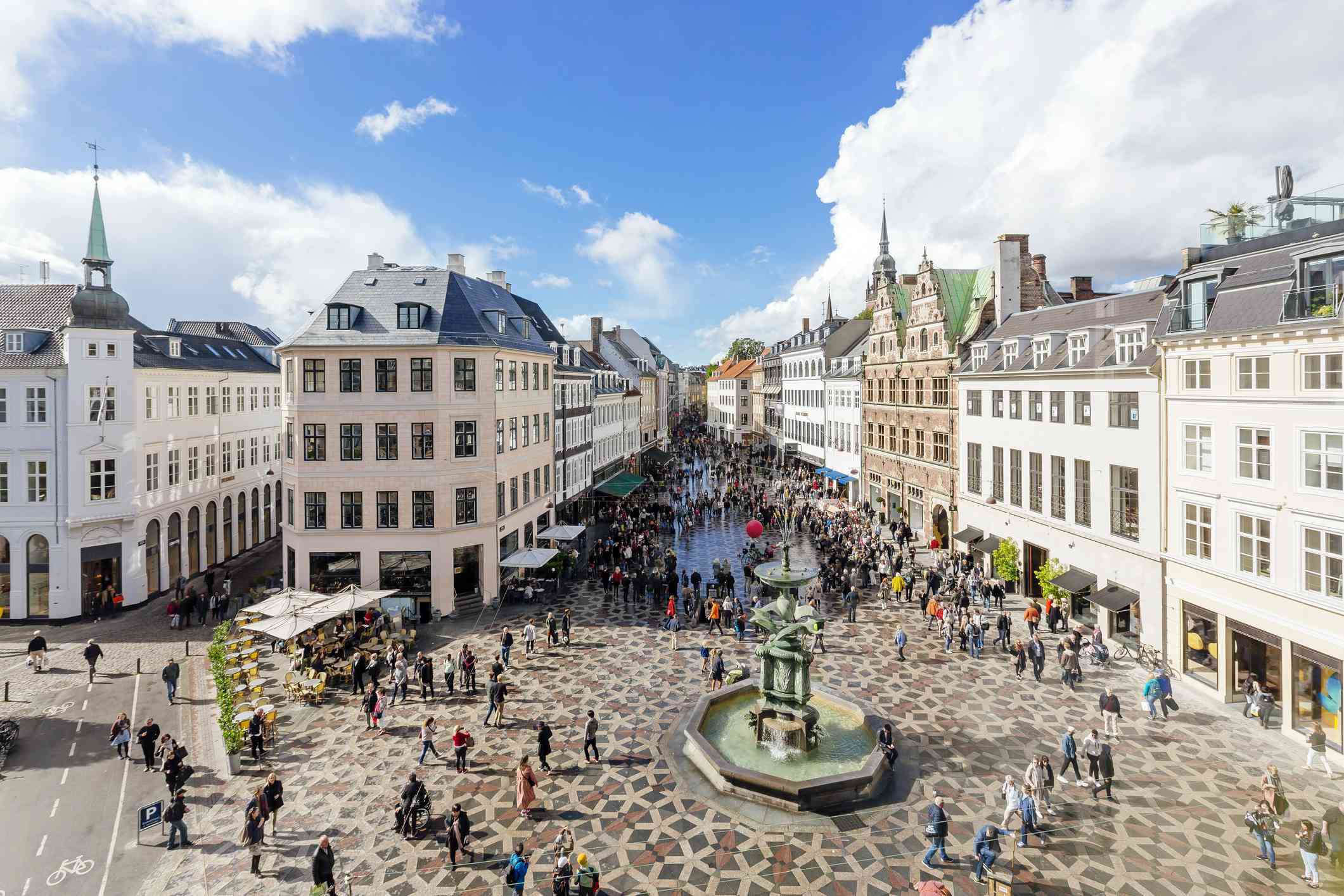 在一个阳光明媚的日子里，人们在蓝天白云下漫步在哥本哈根的阿马格托夫镇广场上，广场两边的建筑中心都有一个喷泉