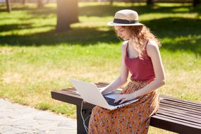 坐在公园长凳上使用笔记本电脑的妇女，旁边有一个太阳能充电器＂width=