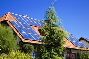 房屋，陡峭的倾斜陶土屋顶覆盖着一系列太阳能电池板，周围有树木和灌木“width=