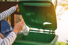 一个人的手在绿色回收箱中扔瓶子。“width=