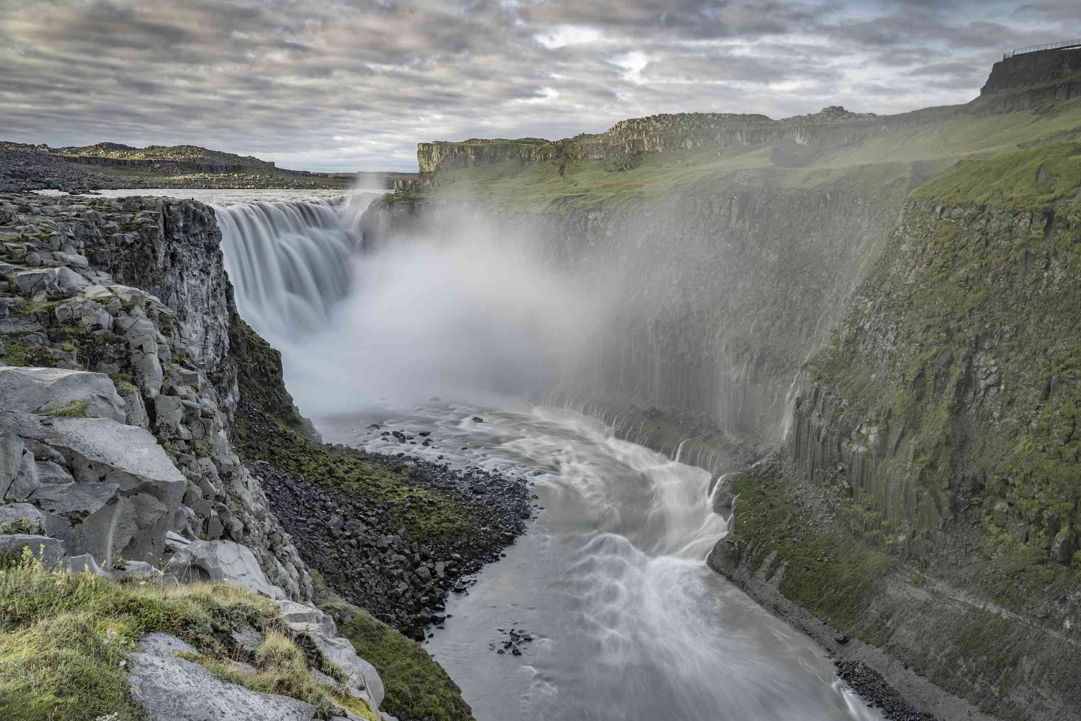 Vatnajokull国家公园中强大的Dettifoss瀑布，水在被绿色覆盖的山脉包围的地面形成一条小溪，瀑布顶部有一点雾＂width=