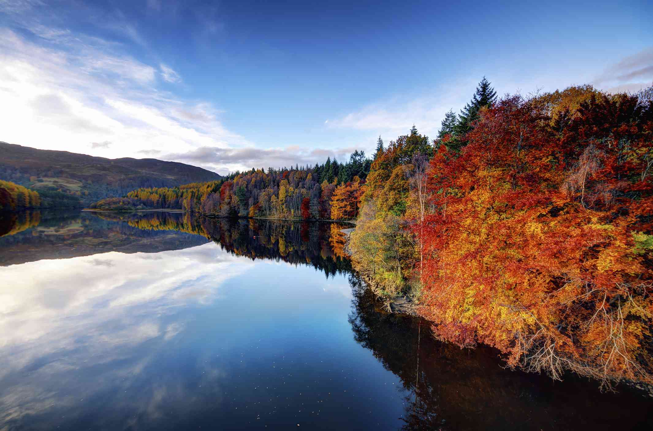 法斯卡利湖的景色，秋天的树木排成红色、金色和绿色的阴影，蓝天和湖面上的白云倒映在湖面上