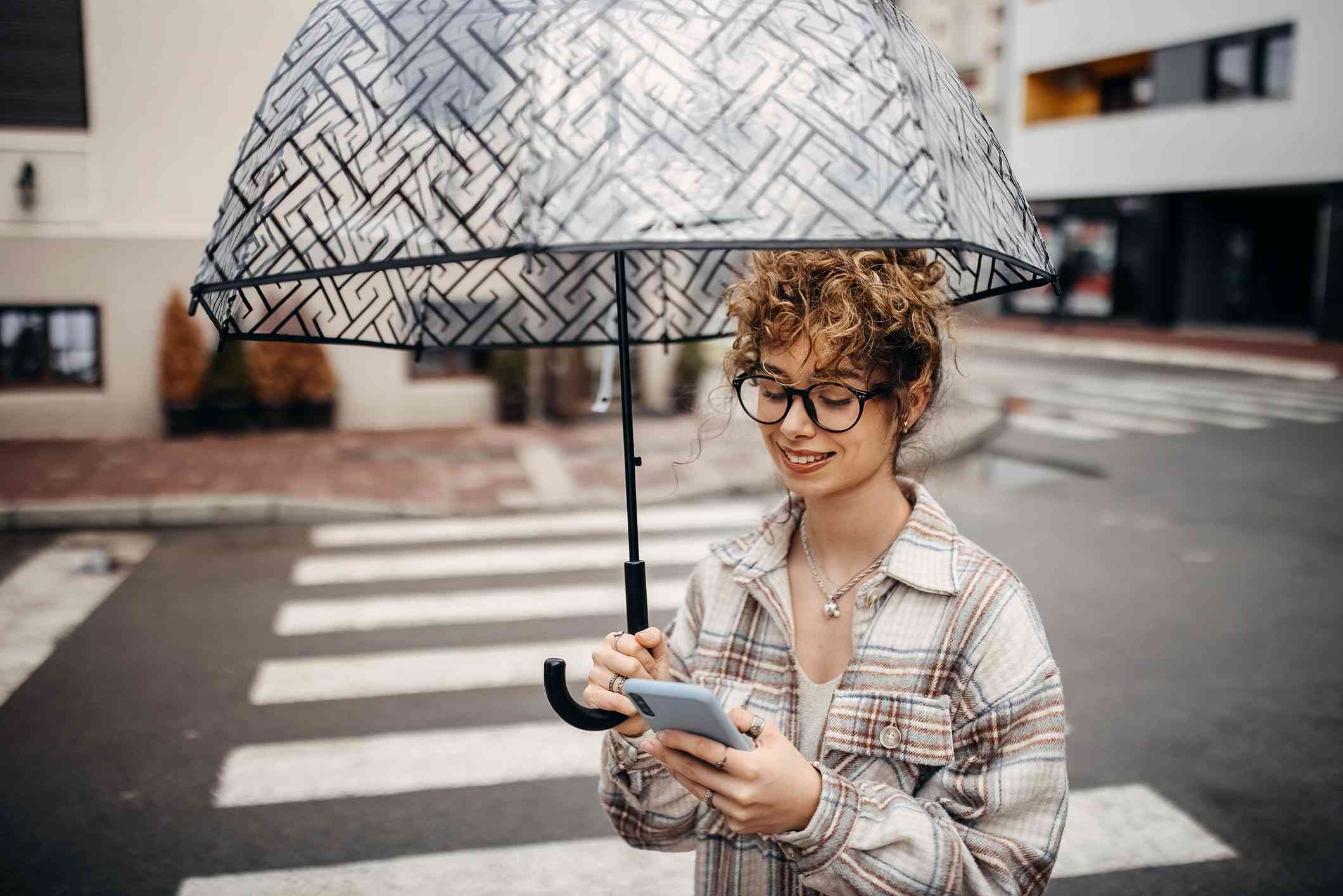 一名年轻女子撑着伞，看着手机穿过马路。