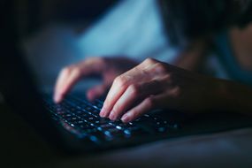 一个年轻女子在黑暗中用笔记本电脑工作到很晚的特写镜头＂width=