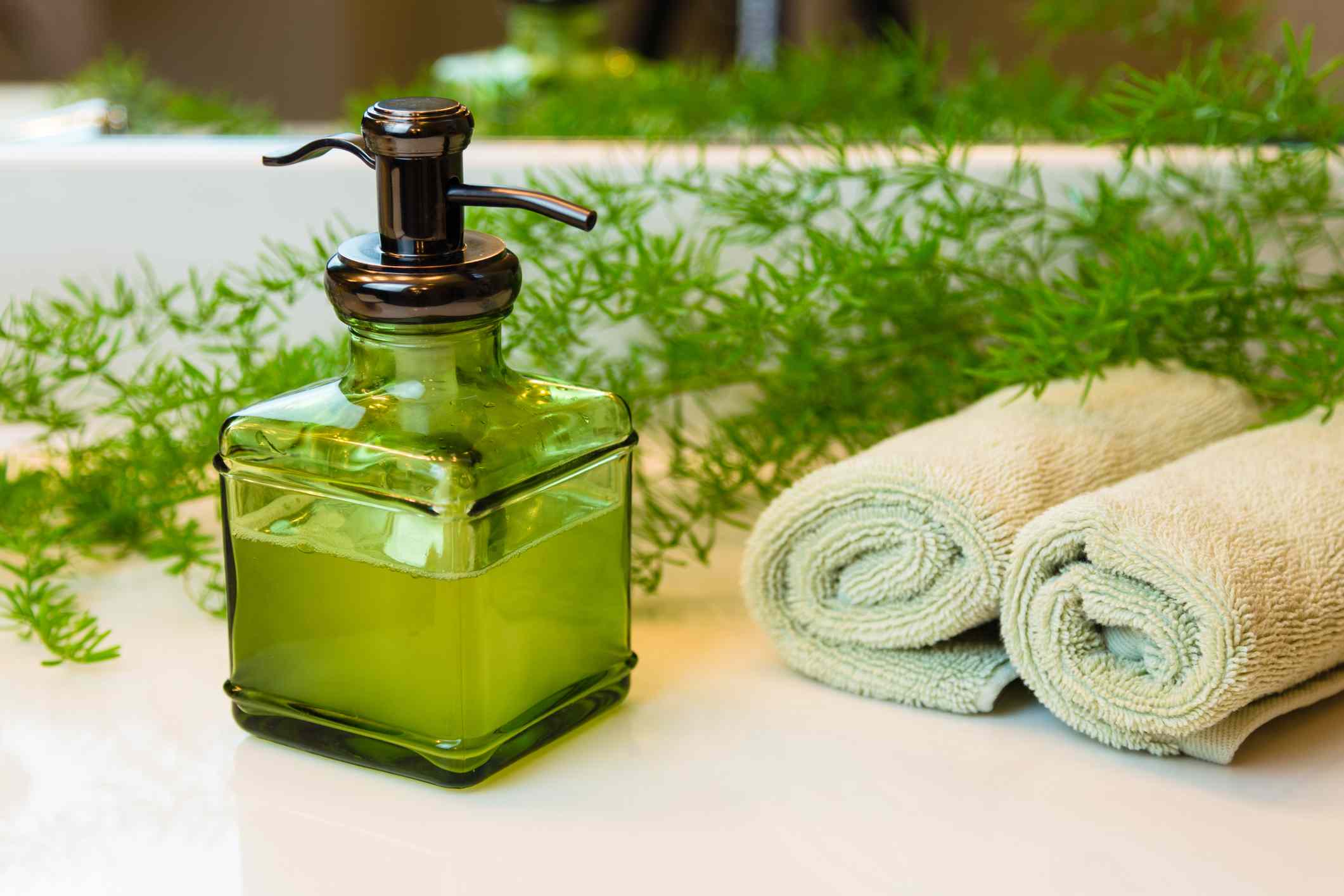 用液体卡斯蒂利亚肥皂泵绿色玻璃瓶。绿色毛巾卷在温泉设置。绿色植物装饰的背景。浴室白色台面