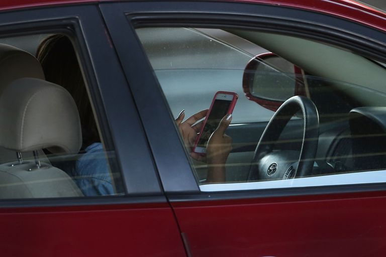 一个人拿着手机在汽车方向盘后面的图像。“class=