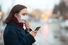 小女孩戴着防护口罩，用智能手机检查空气污染”width=