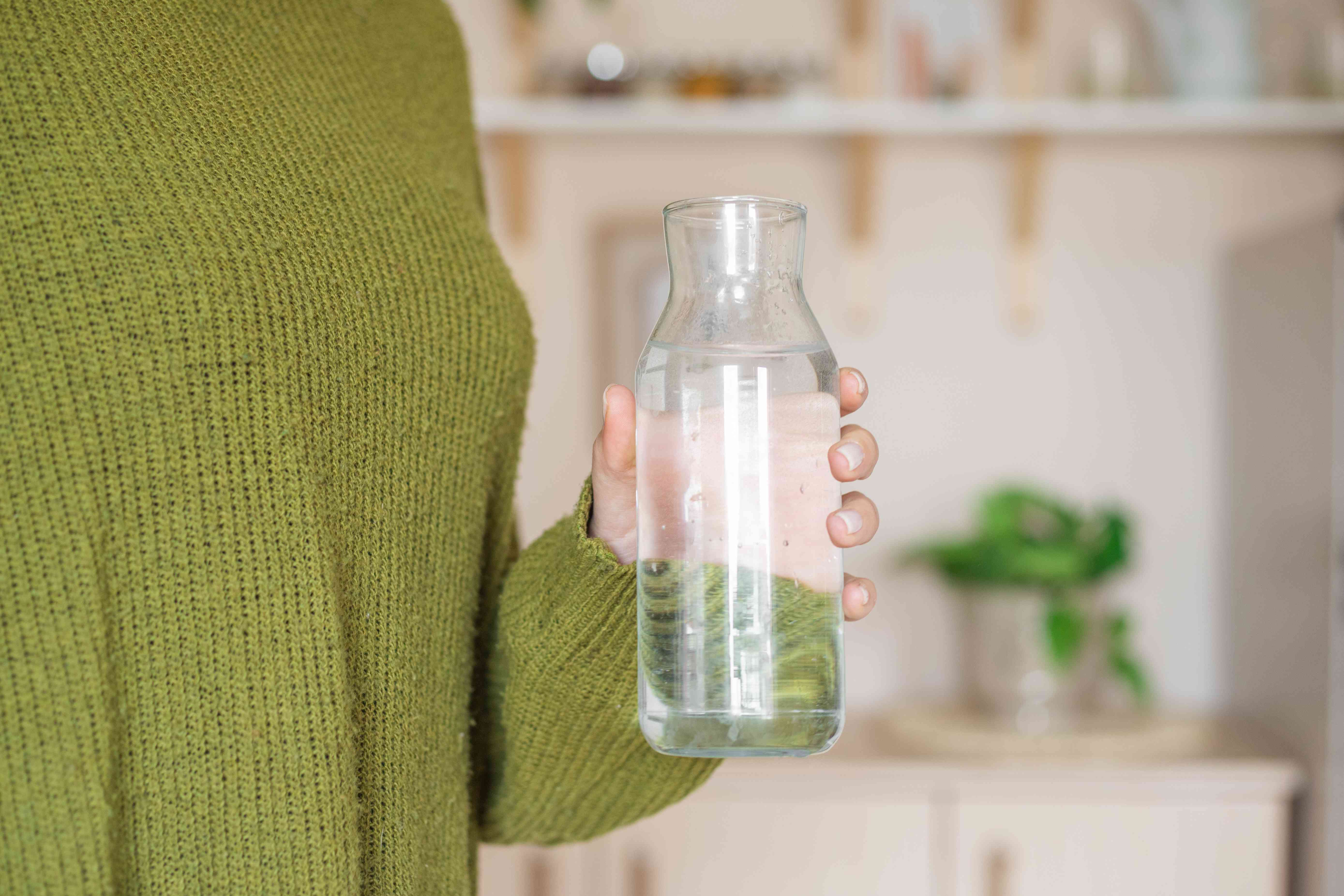 身着橄榄绿毛衣的女子手持透明玻璃水瓶补水