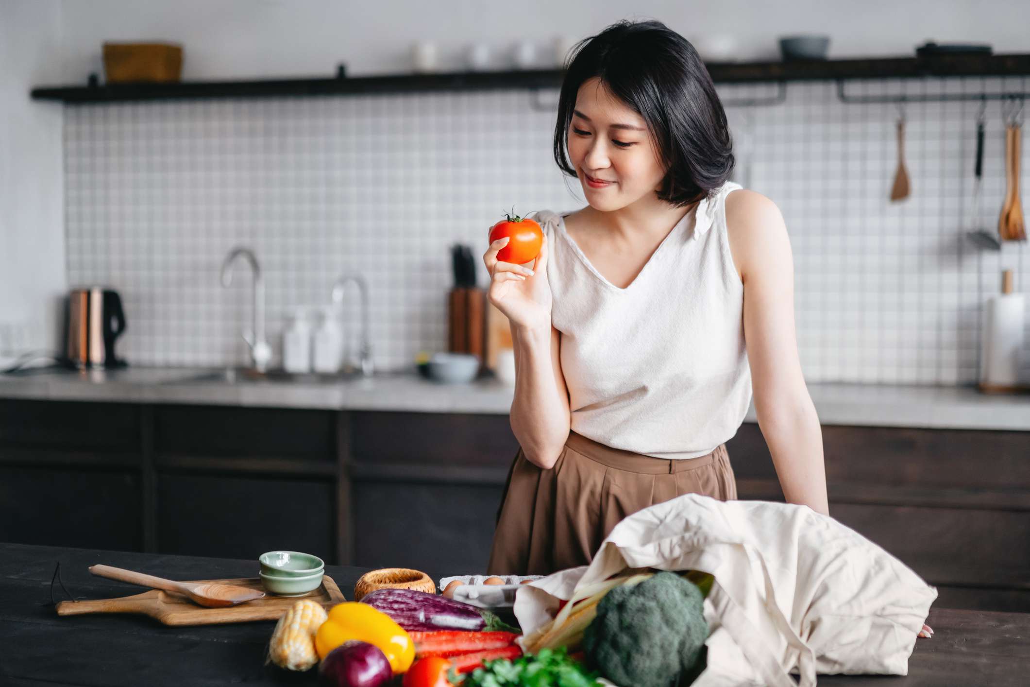 年轻的亚洲女性从杂货店购物回来，从厨房柜台上的可重复使用购物袋里拿出新鲜的水果和蔬菜。她打算用新鲜农产品做一顿健康的饭菜＂width=
