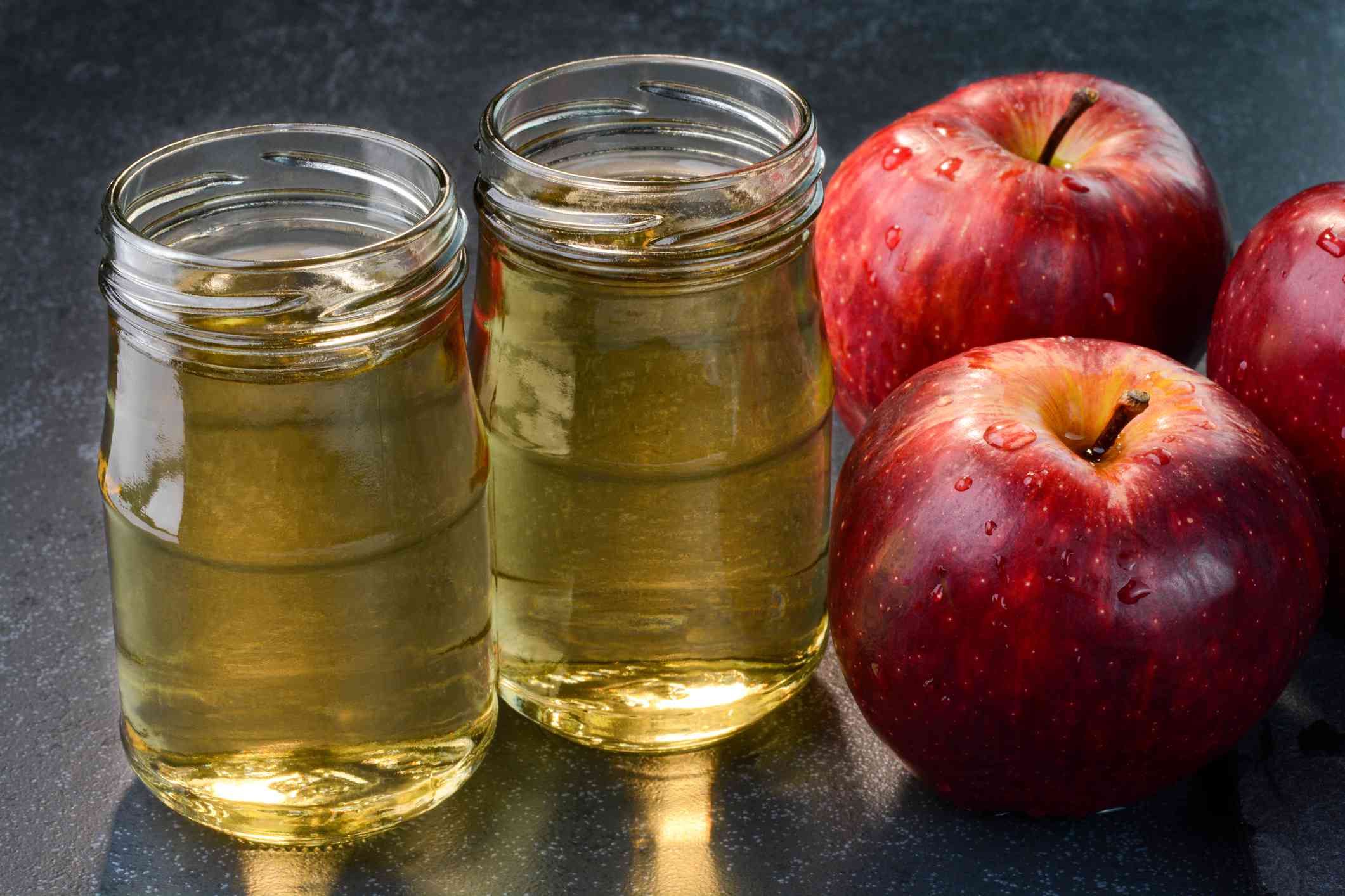 两个小罐子苹果醋坐在接下来的两个三个苹果被水滴覆盖“width=