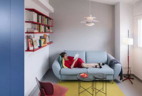 在一间现代公寓里，一个穿着红裙子的女人懒洋洋地躺在蓝色沙发上看书
