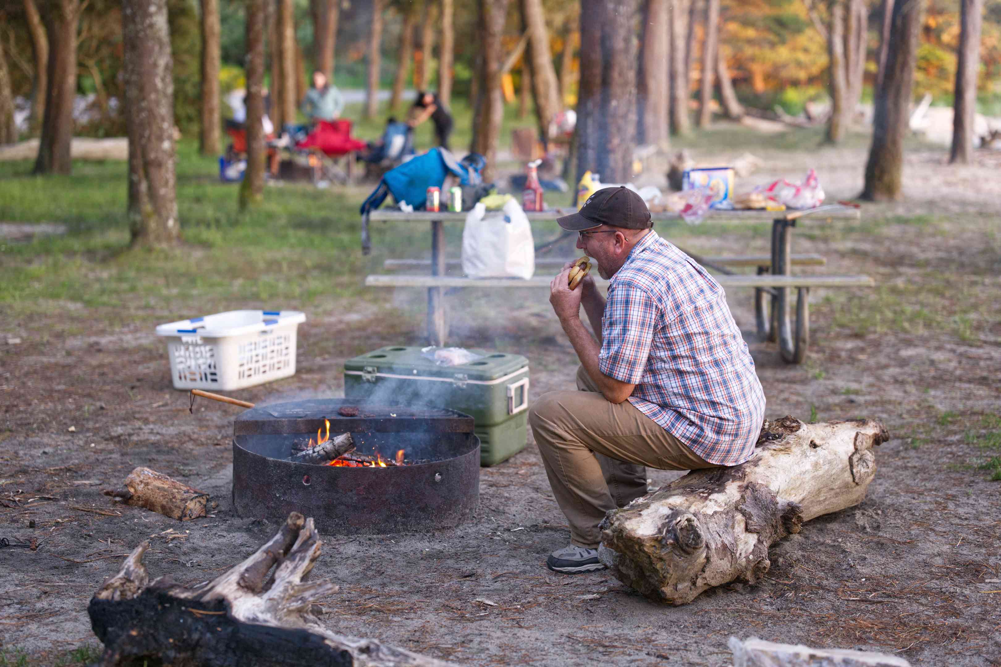 在篝火旁边的露营地，一个家伙坐在原木上，咬了一大口汉堡
