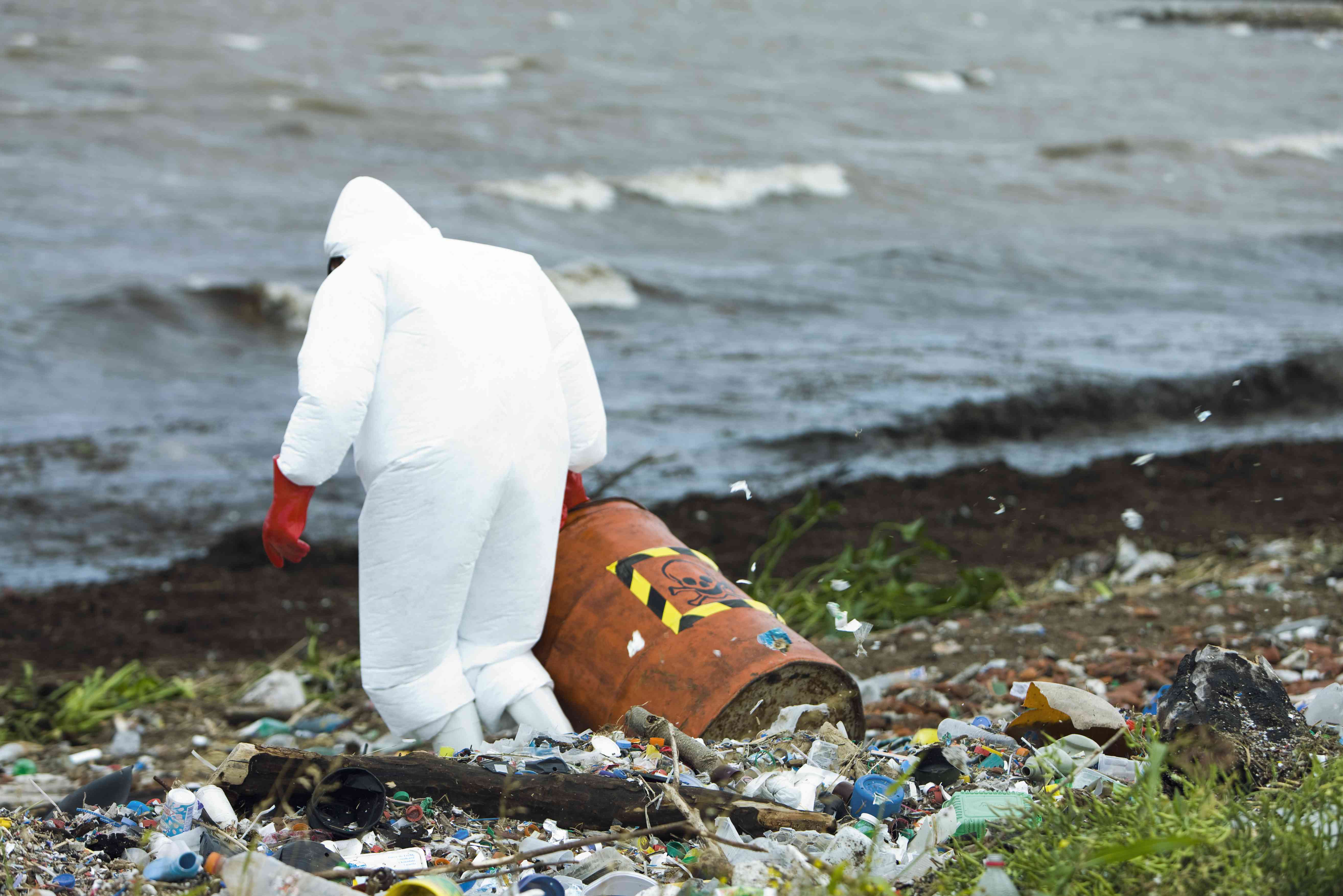 穿着保护套装的人，载有危险废物在污染的海岸上“width=