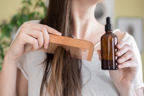 一名女子手持装有辣木油的玻璃瓶，梳理她的长发以保持头发湿润＂width=