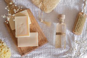 白色华夫饼织物上陈列着肥皂和液体卡斯提尔肥皂，带有木刷和丝瓜络＂width=