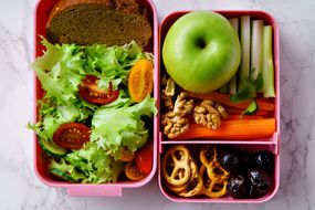 午餐盒有沙拉，面包，苹果，蔬菜，坚果和椒盐卷饼＂width=