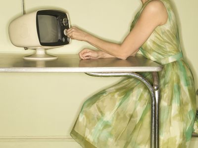 a photo shows a woman wearing a green dress adjusting a small retro tv